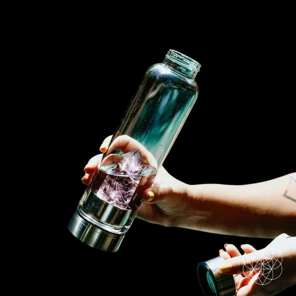 AetherLux™️ glass water bottle with a large amethyst crystal inside, held by hands against a black background.